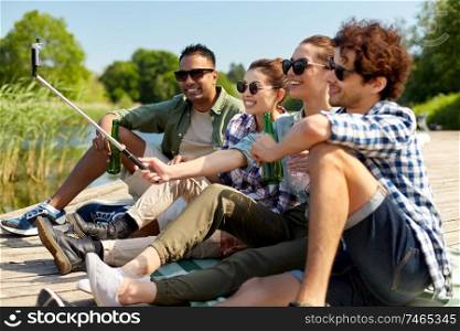 leisure, picnic and technology concept - friends with drinks taking picture by selfie stick on lake pier in summer park. friends with drinks taking selfie on lake pier