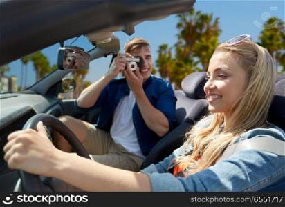 leisure, road trip, travel and people concept - happy couple driving in convertible car and taking picture by film camera over venice beach background in california. man photographing woman driving car by film camera. man photographing woman driving car by film camera