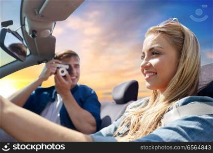 leisure, road trip, travel and people concept - happy couple driving in cabriolet car and taking picture by film camera over evening sky background. happy couple with camera driving in cabriolet car