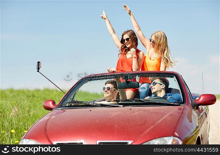 leisure, road trip, travel and people concept - happy friends driving in cabriolet taking picture by smartphone selfie stick at country
