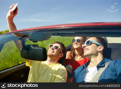 leisure, road trip, travel and people concept - happy friends driving in cabriolet taking selfie by smartphone outdoors