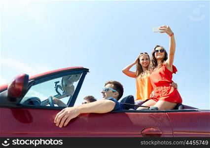 leisure, road trip, travel and people concept - happy friends driving in cabriolet taking selfie by smartphone outdoors