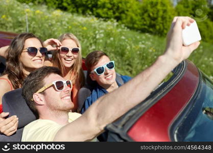 leisure, road trip, travel and people concept - happy friends driving in cabriolet taking selfie by smartphone at country