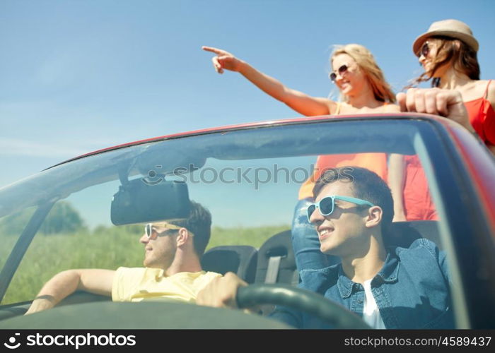 leisure, road trip, travel and people concept - happy friends driving in cabriolet car along country road and pointing finger to something