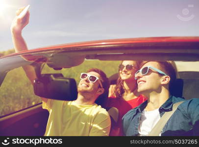 leisure, road trip, travel and people concept - happy friends driving in cabriolet taking selfie by smartphone outdoors. friends driving in cabriolet car and taking selfie