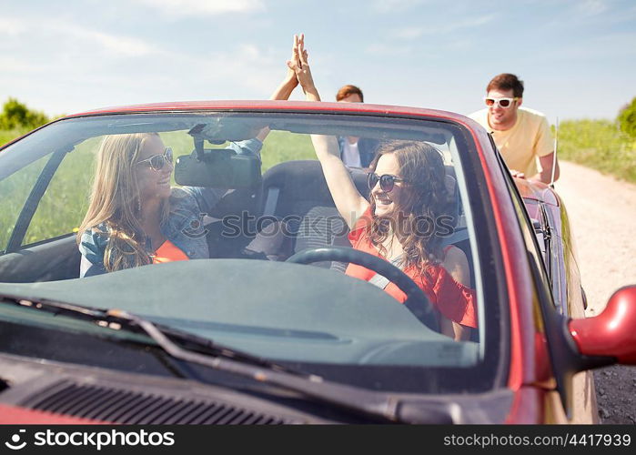 leisure, road trip, travel and people concept - happy friends pushing broken cabriolet car along country road