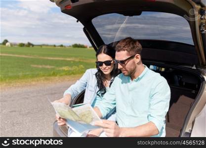 leisure, road trip, travel and people concept - happy man and woman searching location on map sitting on trunk of hatchback car outdoors