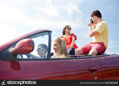 leisure, road trip, travel, summer holidays and people concept - happy friends driving in cabriolet car and taking picture by film camera