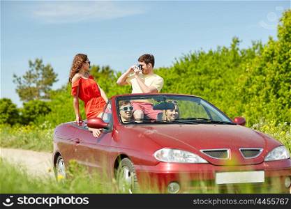 leisure, road trip, travel, summer holidays and people concept - happy friends driving in cabriolet car and taking picture by film camera