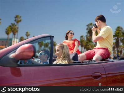 leisure, road trip, travel, summer holidays and people concept - happy friends driving in convertible car and taking picture by film camera over venice beach background in california. friends with camera driving in convertible car
