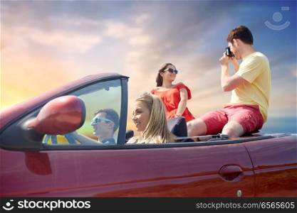 leisure, road trip, travel, summer holidays and people concept - happy friends driving in convertible car and taking picture by film camera over sky background. happy friends with camera driving in car