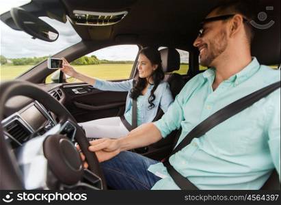 leisure, road trip, travel, technology and people concept - happy man and woman driving in car and taking selfie by smartphone