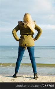 Leisure, spending free time outside, healthy walks concept. Woman wearing warm jacket relaxing on beach near sea, cold sunny day, back view. Woman relaxing on beach, cold day