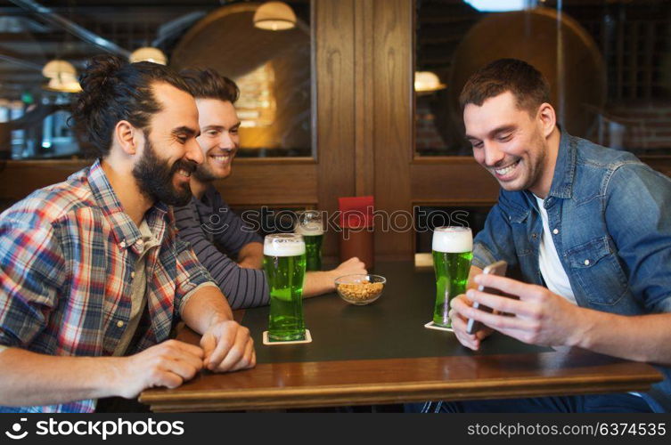 leisure, st patricks day and technology concept - happy male friends with smartphone drinking green beer at bar or pub. friends with smartphone drinking green beer at pub