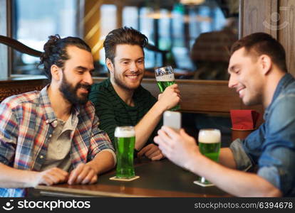 leisure, st patricks day and technology concept - happy male friends with smartphone drinking green beer at bar or pub. friends with smartphone drinking green beer at pub