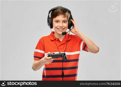 leisure, technology and children concept - smiling boy in headphones with microphone and gamepad playing video game over grey background. boy in headphones with gamepad playing video game