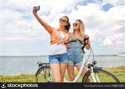leisure, technology and friendship concept - happy smiling teenage girls or friends with bicycle taking selfie by smartphone at seaside in summer. teenage girls with bicycle taking selfie in summer