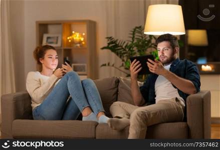 leisure, technology and internet addiction concept - couple with tablet computer and smartphone at home in evening. couple with tablet computer and smartphone at home