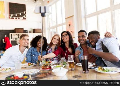 leisure, technology and people concept - group of happy international friends eating and taking picture by smartphone selfie stick at restaurant. friends eating and taking selfie at restaurant