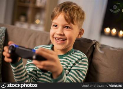 leisure, technology and people concept - happy little boy with gamepad playing video game at home in evening. little boy with gamepad playing video game at home
