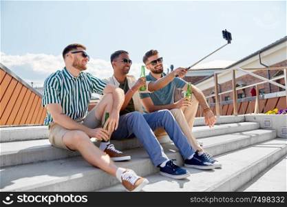 leisure, technology and people concept - happy male friends taking picture by smartphone on selfie stick and drinking beer on street in summer. men drinking beer and taking selfie by smartphone