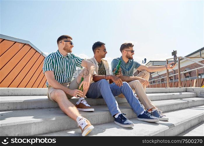 leisure, technology and people concept - happy male friends taking selfie by smartphone on tripod and drinking beer on street in summer. men drinking beer and taking selfie by smartphone