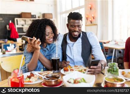 leisure, technology and people concept - happy man and woman with smartphones eating at restaurant. happy man and woman with smartphones at restaurant