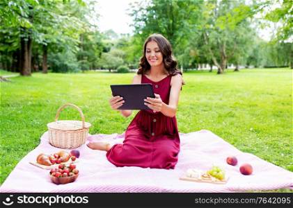 leisure, technology and people concept - happy smiling woman with tablet computer, picnic basket and food sitting on blanket at summer park. happy woman with tablet computer on picnic at park