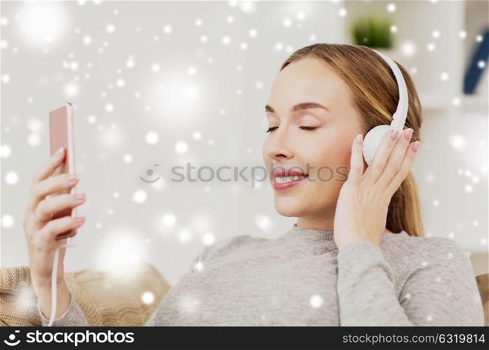 leisure, technology and people concept - smiling woman with smartphone and headphones listening to music at home over snow. woman with smartphone and headphones at home