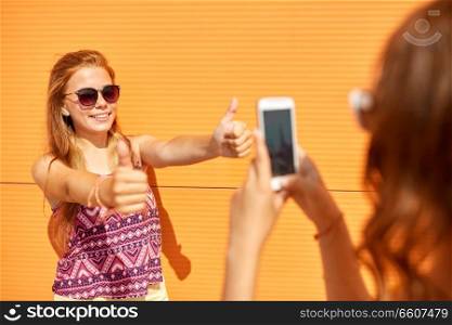 leisure, technology and people concept - teenage girl taking picture of her smiling friend by smartphone outdoors in summer. teenage girl photographing friend by smartphone