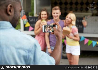 leisure, technology and people concept - young man taking picture of his happy friends eating hamburgers and wok at food truck. man taking picture of friends eating at food truck