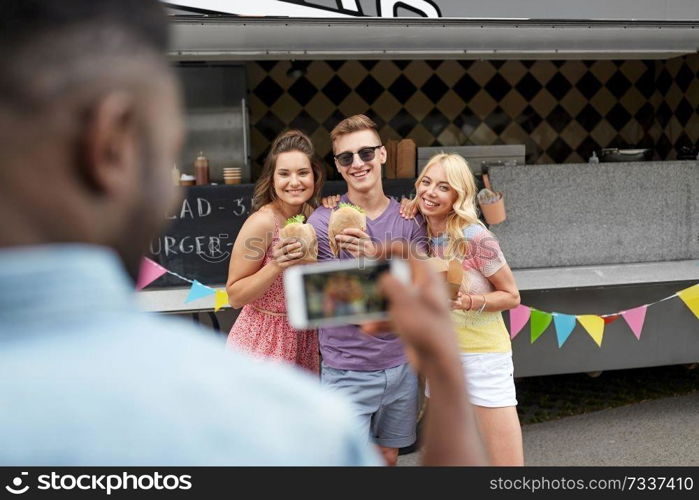 leisure, technology and people concept - young man taking picture of his happy friends eating hamburgers and wok at food truck. man taking picture of friends eating at food truck