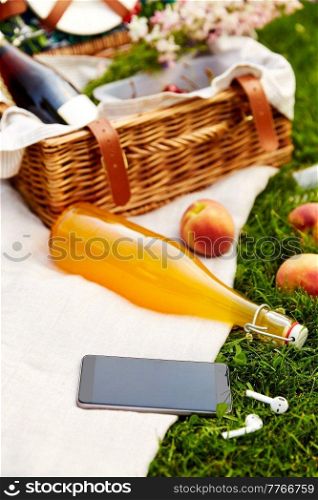 leisure, technology and summer concept - close up of picnic basket, bottle of fruit juice and smartphone with earphones on grass. phone, earphones, picnic basket and juice bottle