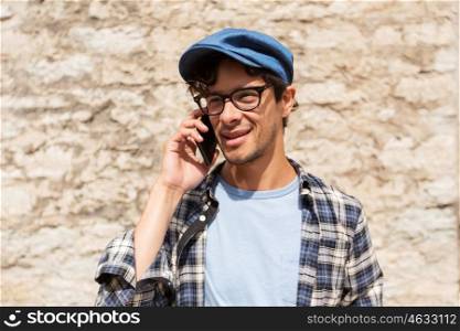 leisure, technology, communication and people concept - close up of hipster man in hat and glasses calling on smartphone at street wall