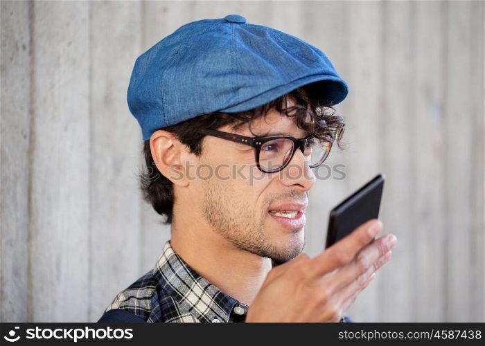 leisure, technology, communication and people concept - smiling hipster man using voice command recorder or calling on smartphone at street wall. man recording voice or calling on smartphone