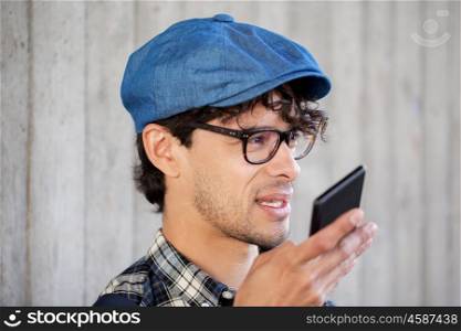 leisure, technology, communication and people concept - smiling hipster man using voice command recorder or calling on smartphone at street wall. man recording voice or calling on smartphone
