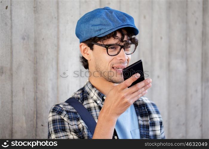 leisure, technology, communication and people concept - smiling hipster man using voice command recorder or calling on smartphone at street wall