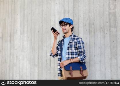 leisure, technology, communication and people concept - smiling hipster man with shoulder bag using voice command recorder or calling on smartphone at street wall
