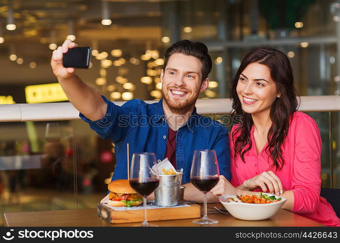 leisure, technology, date, people and holidays concept - happy couple having dinner and taking selfie by smartphone at restaurant