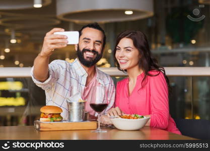 leisure, technology, date, people and holidays concept - happy couple having dinner and taking selfie by smartphone at restaurant