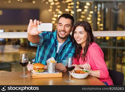 leisure, technology, date, people and holidays concept - happy couple having dinner and taking selfie by smartphone at restaurant
