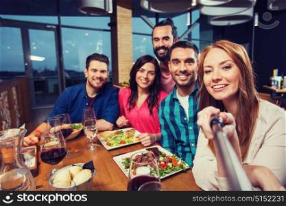 leisure, technology, friendship, people and holidays concept - happy friends having dinner and taking picture by selfie stick at restaurant. friends picturing by selfie stick at restaurant