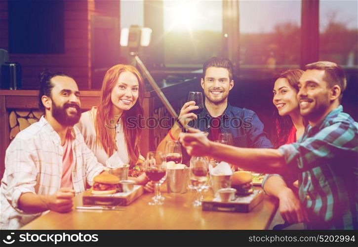 leisure, technology, friendship, people and holidays concept - happy friends having dinner and taking picture by smartphone selfie stick at restaurant. friends taking selfie by smartphone at restaurant