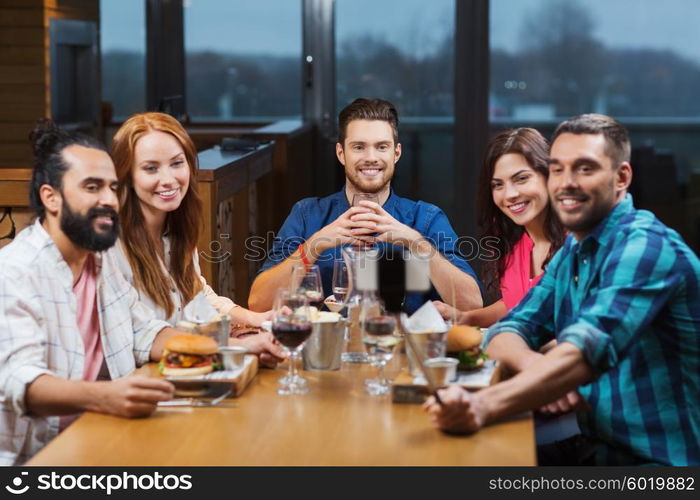 leisure, technology, friendship, people and holidays concept - happy friends having dinner and taking picture by smartphone selfie stick at restaurant