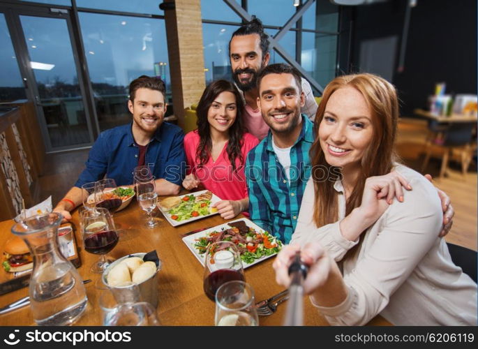 leisure, technology, friendship, people and holidays concept - happy friends having dinner and taking picture by smartphone selfie stick at restaurant