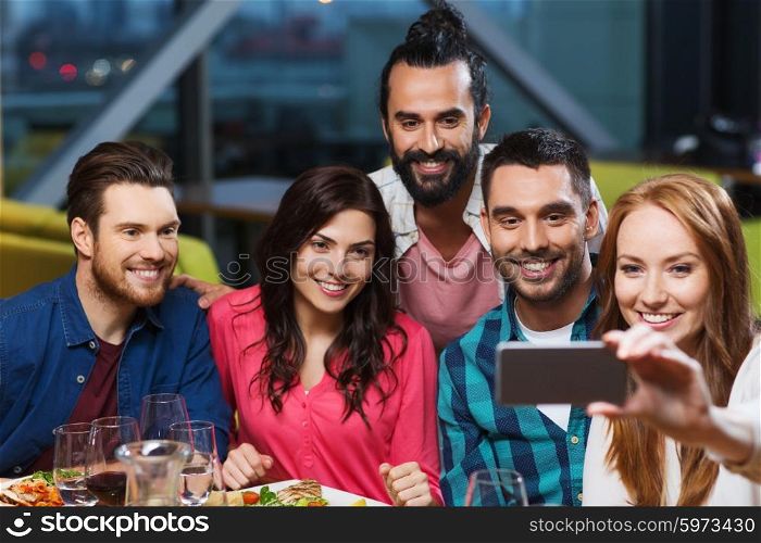 leisure,technology, friendship, people and holidays concept - happy friends having dinner and taking selfie by smartphone at restaurant