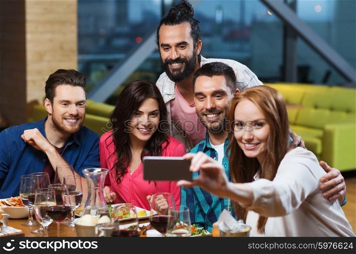 leisure,technology, friendship, people and holidays concept - happy friends having dinner and taking selfie by smartphone at restaurant