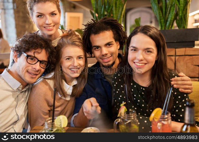 leisure, technology, friendship, people and holidays concept - happy friends with drinks and taking picture by smartphone selfie stick at bar or cafe