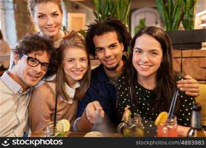 leisure, technology, friendship, people and holidays concept - happy friends with drinks and taking picture by smartphone selfie stick at bar or cafe