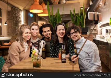 leisure, technology, friendship, people and holidays concept - happy friends with drinks and taking picture by smartphone selfie stick at bar or cafe. friends taking selfie by smartphone at bar or cafe